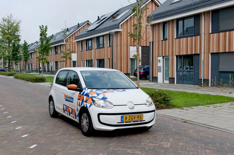 Witte auto, die de kleurelementen en het logo van De Woonplaats bevat, rijdt door een straat met huizen